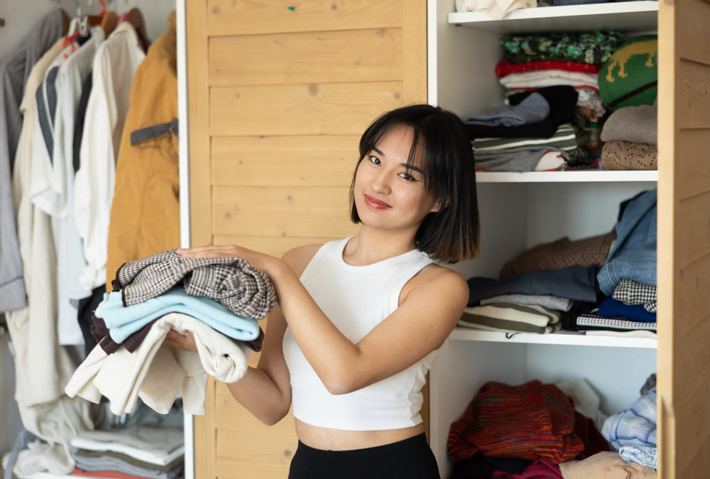 Happy young Asian girl holding some items of clothing by the wardrobe