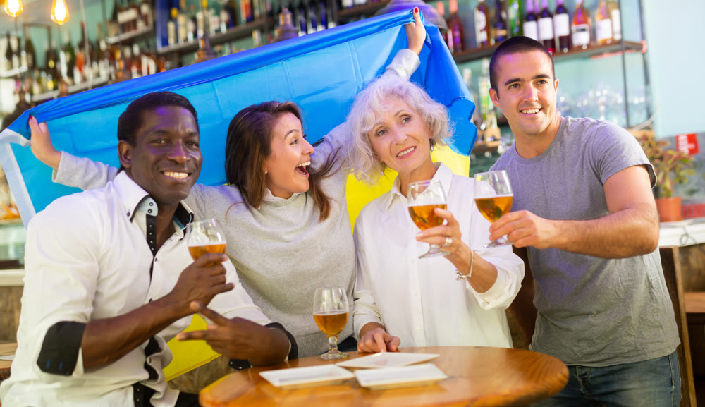 Excited male and female football fans celebrating championship win, waving flag of Ukraine in pub