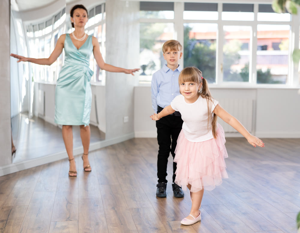 Girl rehearsing elegant curtsy with boy dance partner and female teacher