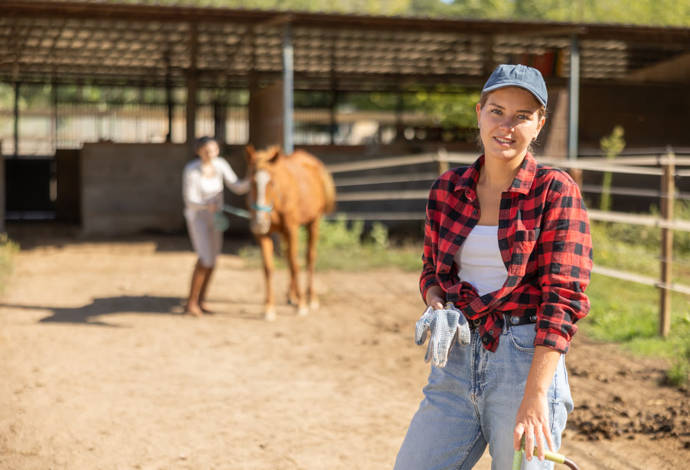 Positive female stable worker stands near wooden fence of paddock and watches horse.