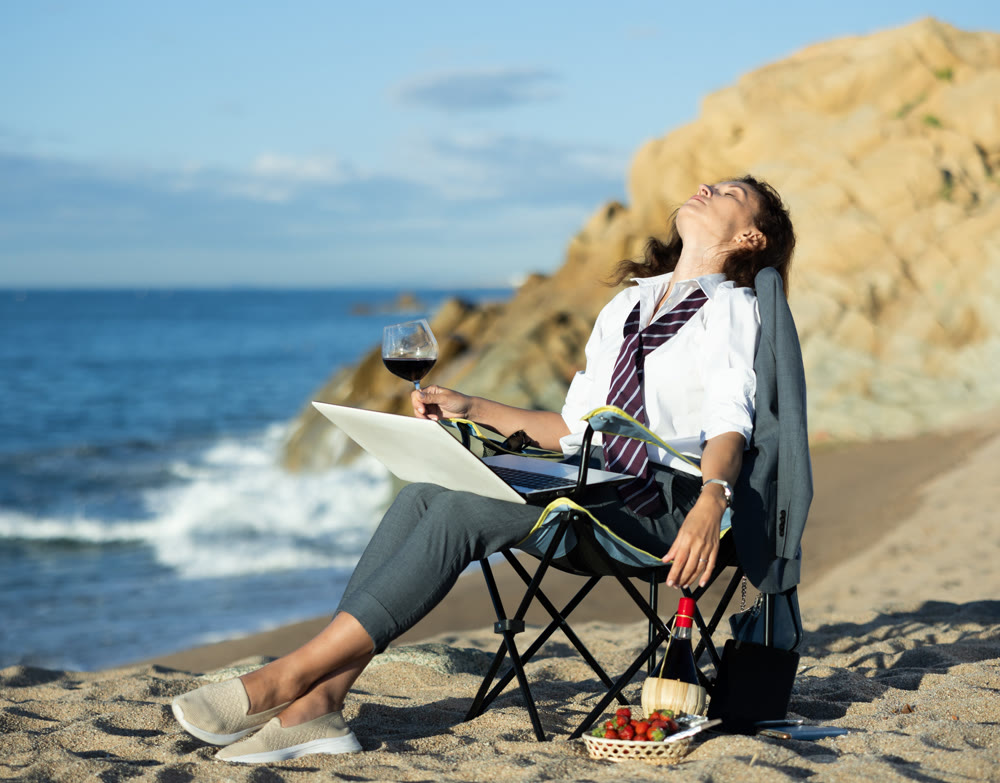 Stylish woman in a suit works on her laptop by the ocean and drinking wine