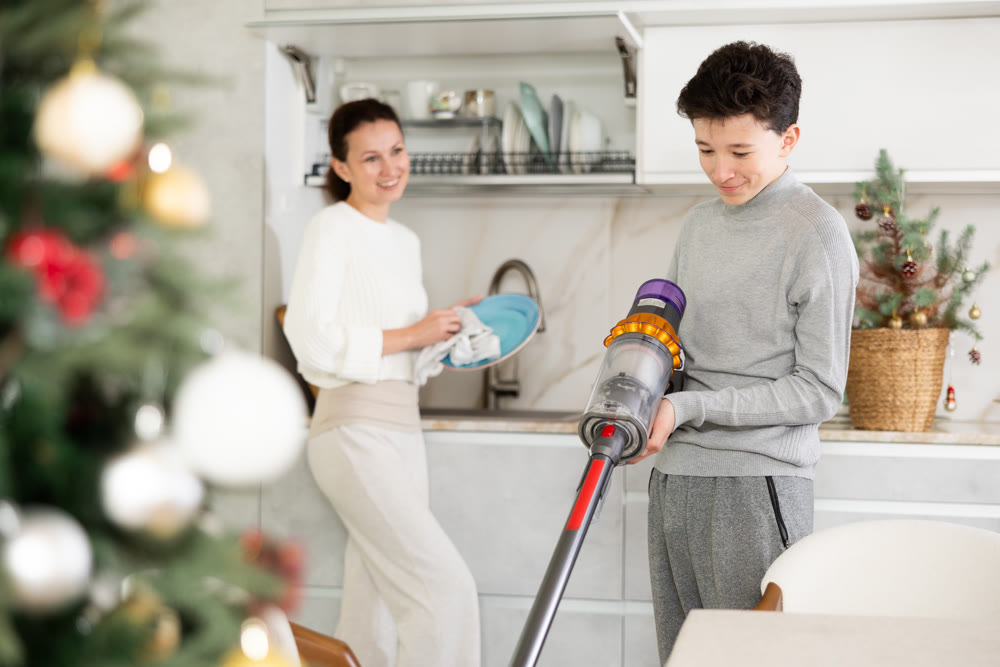 Mother and her teenage son are preparing to celebrate Christmas at home - they are cleaning kitchen, vacuuming floor and washing dishes