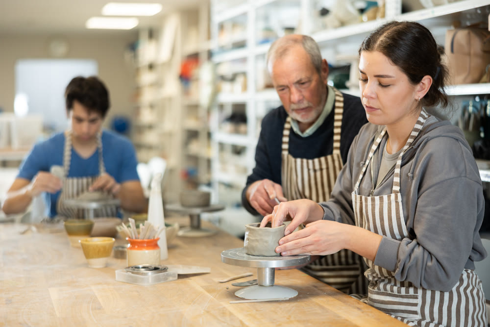 Elderly man teacher shows girl how to sculpt ceramic product from raw clay in workshop
