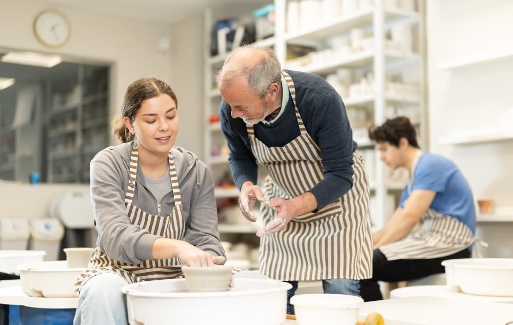 Eldery man helping young woman shape pottery on wheel