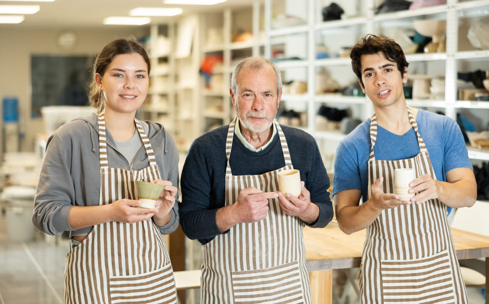 Three positive potters stand in pottery workshop and show created work, finished decorative jug