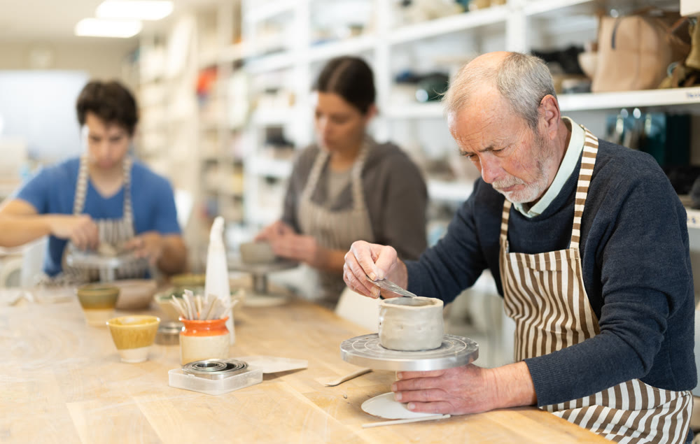 Mature man at table molds cup from wet clay in pottery workshop