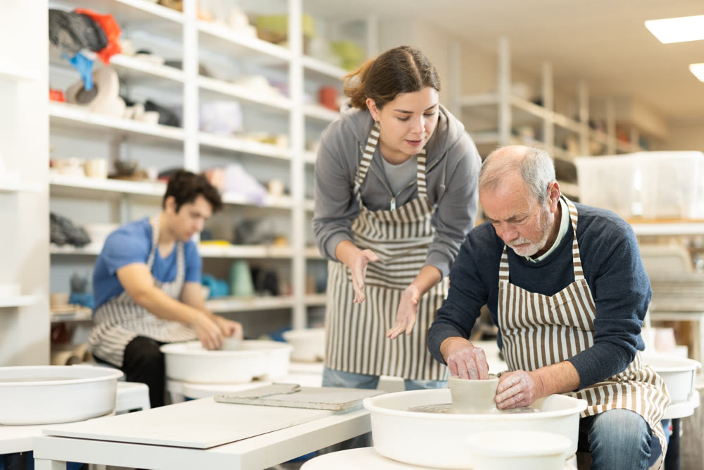 Girl help to mature man working on potters wheel in pottery workshop