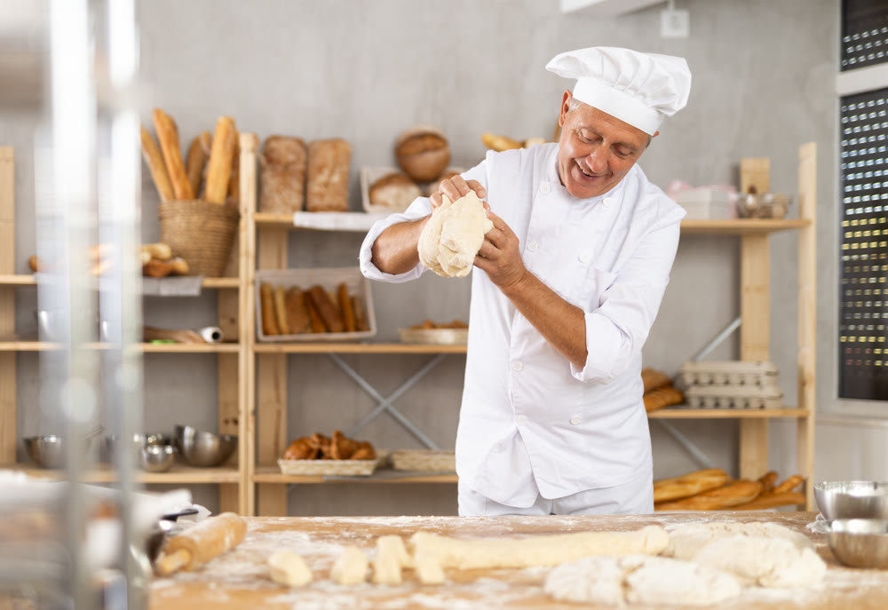 Elderly male baker in uniform prepares raw dough, kneads it with hands