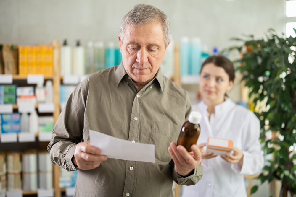 Elderly male customer chooses cough syrup in modern pharmacy. Female pharmacist helps with choosing medicine