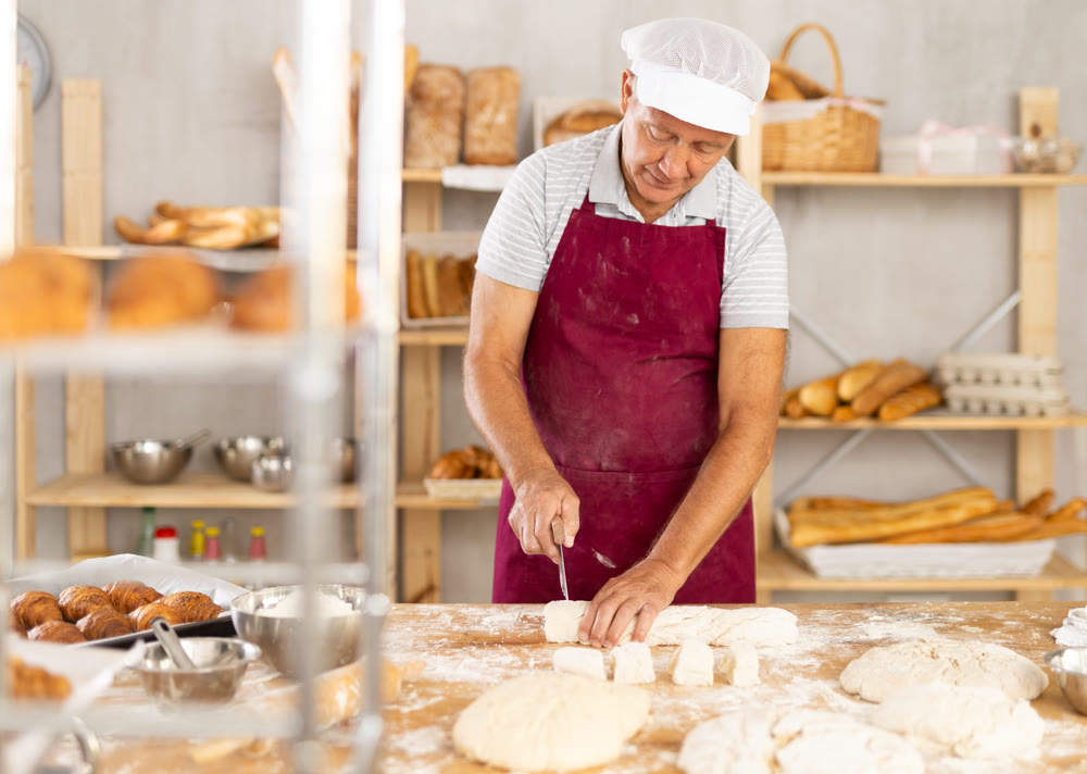 Elderly male baker cutting dough with scraper