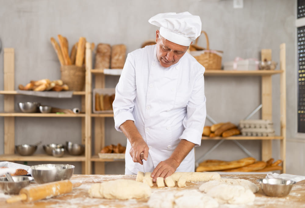 Baker in uniform cutting raw dough with knife to make croissants or buns