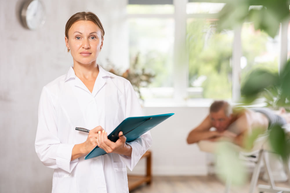 Positive middle-aged masseuse posing with folder in hand in the therapy room