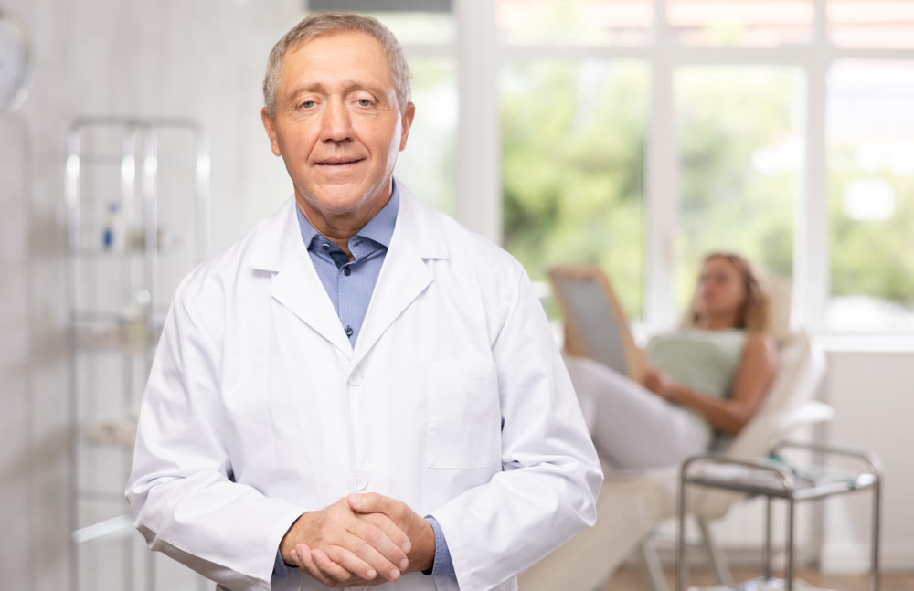 Elderly doctor standing in clinic office with patient lying on couch