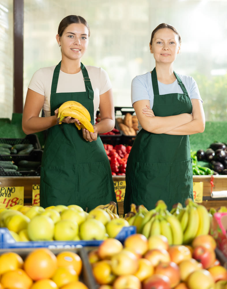 Friendly woman owner of fruit-vegetable shop standing in trading floor with girl assistant