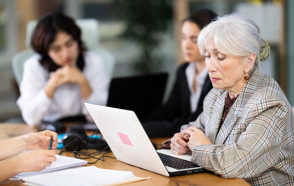 Tired mature woman is sitting at computer at work in crowded office