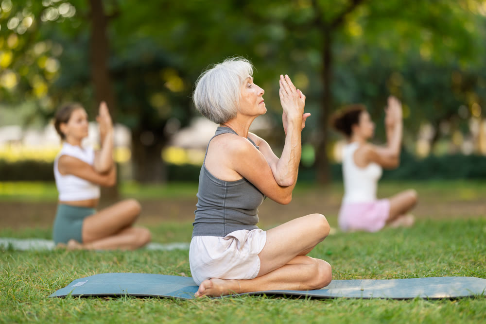 Elderly woman performing Gomukhasana Garudasana during outdoor group yoga training