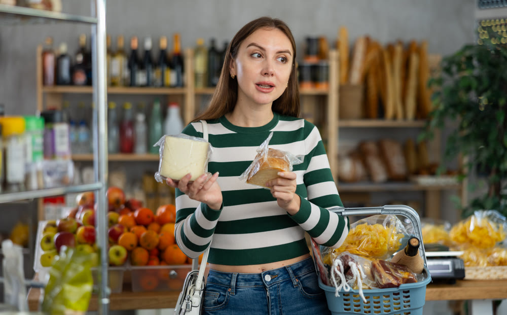 Smiling young girl with shopping basket trying to make choice holding two sorts of cheese in grocery store