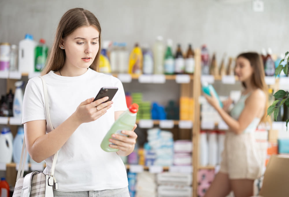 Young female shopper scans QR code on label of dishwashing using her smartphone, while in background shoppers browse products
