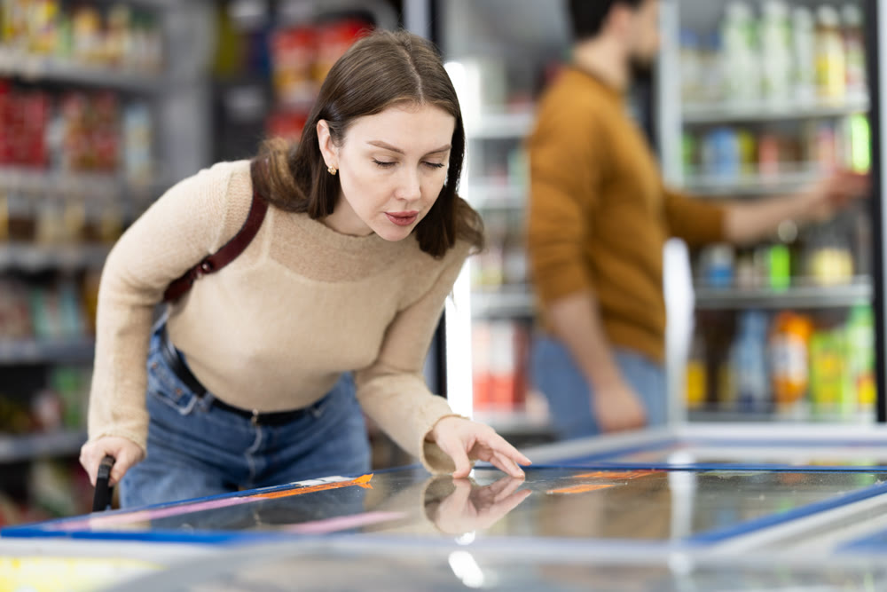 Woman standing near refrigerated display cases choosing frozen food in supermarket