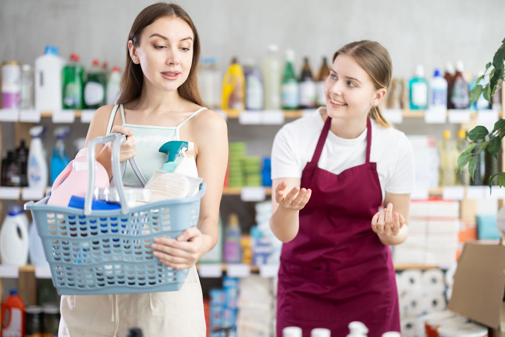 Female salesperson and customer with shopping basket choosing goods together in supermarket