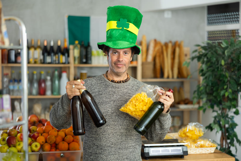 Adult positive man buyer in green costume and hat, shopping bottles of festive beer in store for St. Patrick's Day