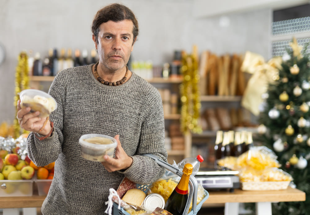 Positive adult man with shopping cart picking out fresh cheese to buy in grocery store for christmas table