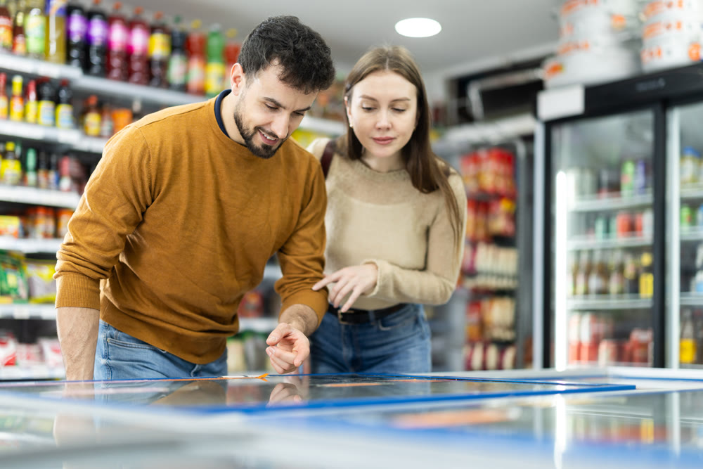 Positive young couple choosing cooled food product standing at fridge in grocery store
