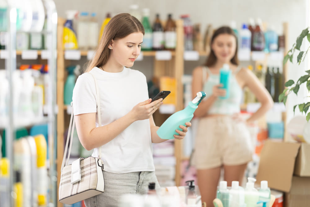Young female shopper scans QR code on label of spray glass cleaner for windows using her smartphone, while in background shoppers browse products