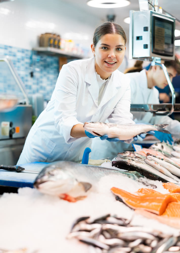 Female seller in apron standing near counter offering fresh fish hake to buyer
