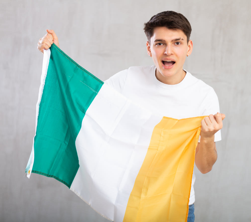 Enthusiastic young man holding flag of Ireland against unicoloured background