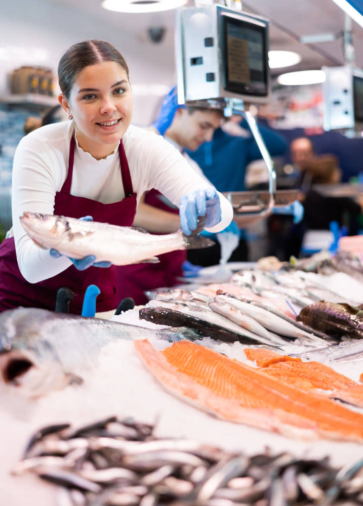 Female seller in apron standing near counter offering fresh fish hake to buyer