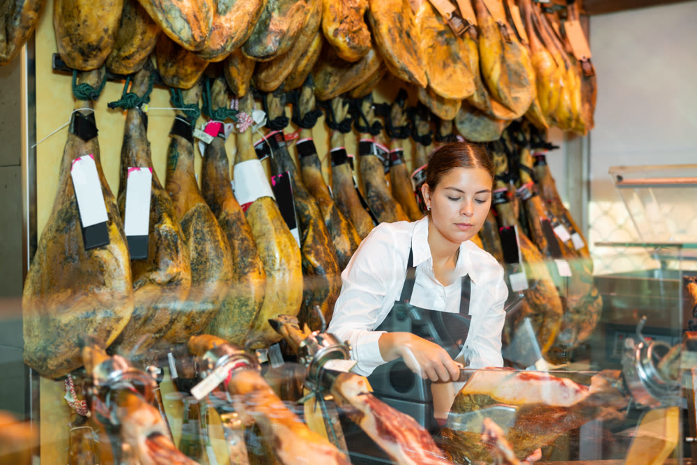 Young saleswoman in black apron slicing Iberian jamon at counter in butcher shop