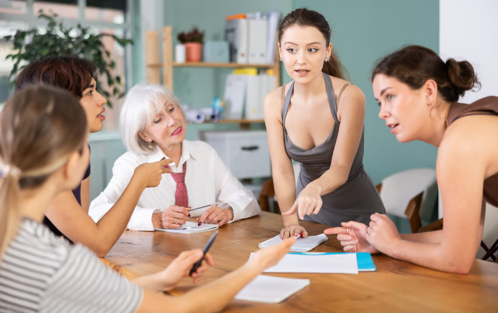 Positive young girl office worker standing with pen in hands near table where sitting colleagues with papers and talking in office