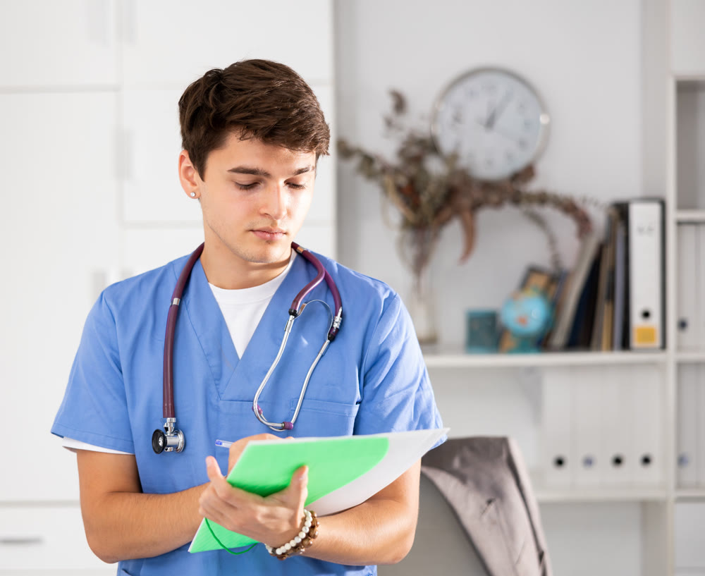 Positive male doctor who works at the clinic writes important notes on folder of documents while