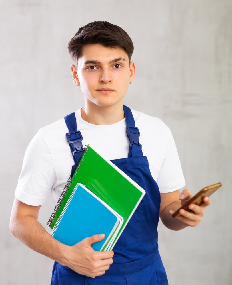 Young male builder in protective overalls with notebooks in his hands writes a message on smartphone screen