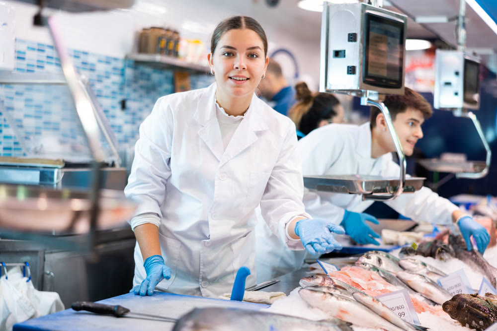 Friendly woman seller in a white coat and gloves invites you to buy fresh fish in supermarket
