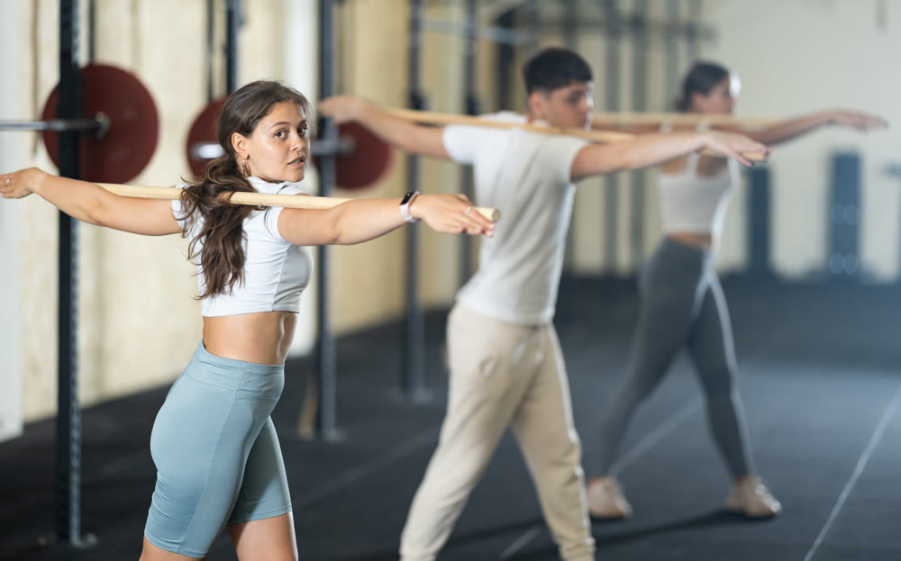 Slender girl, together with other athletes, does a warm-up using a long stick in gym