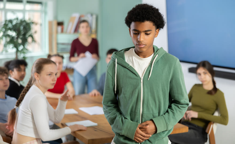 Sad African-American boy stands in study room, group of classmates behind him.
