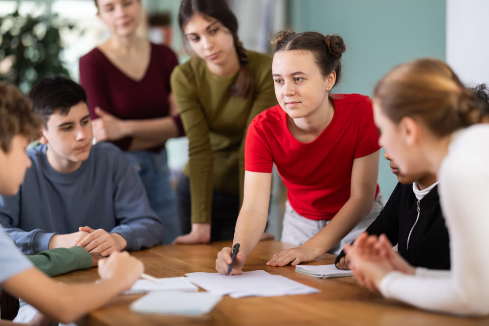 At round table meeting, teen participants discuss problem and try to find solution, teacher watching