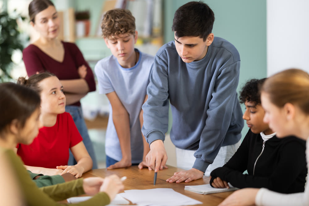 Teenage students solving problem on paper while teacher watches behind