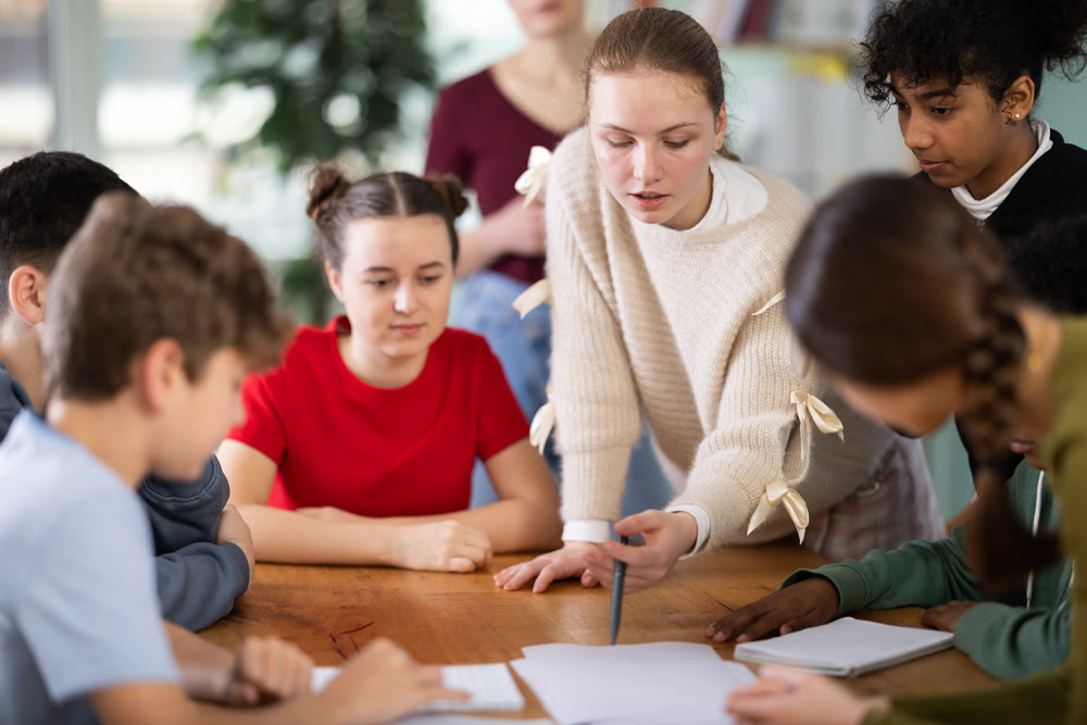 At round table meeting, teen participants discuss problem and try to find solution, teacher watching
