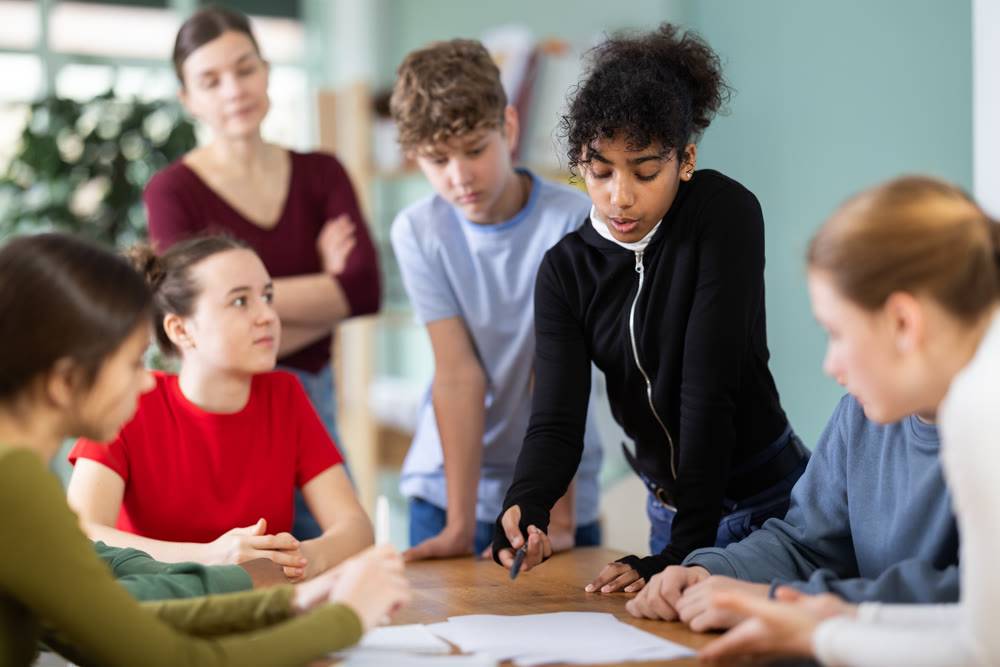 At round table meeting, teen participants discuss problem and try to find solution, teacher watching