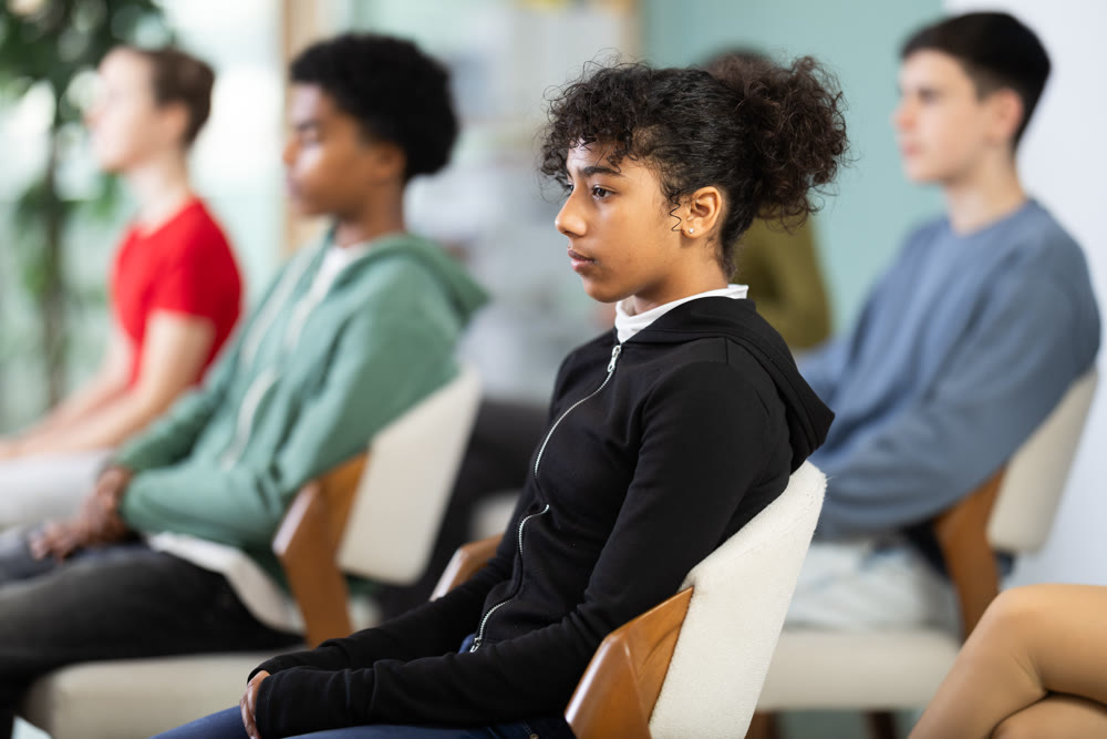 Teenage girl listening to teacher while sitting with classmates in classroom