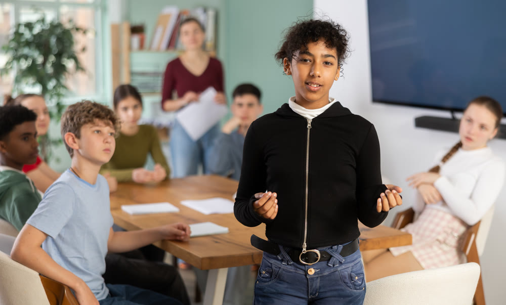 Glad teenage girl standing with her back to classmates sitting at table