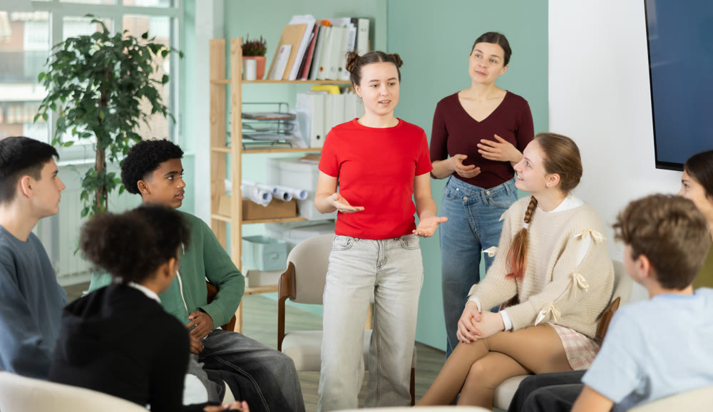 Teen group sit in circle and listen to girl classmate speaking, teacher watching.