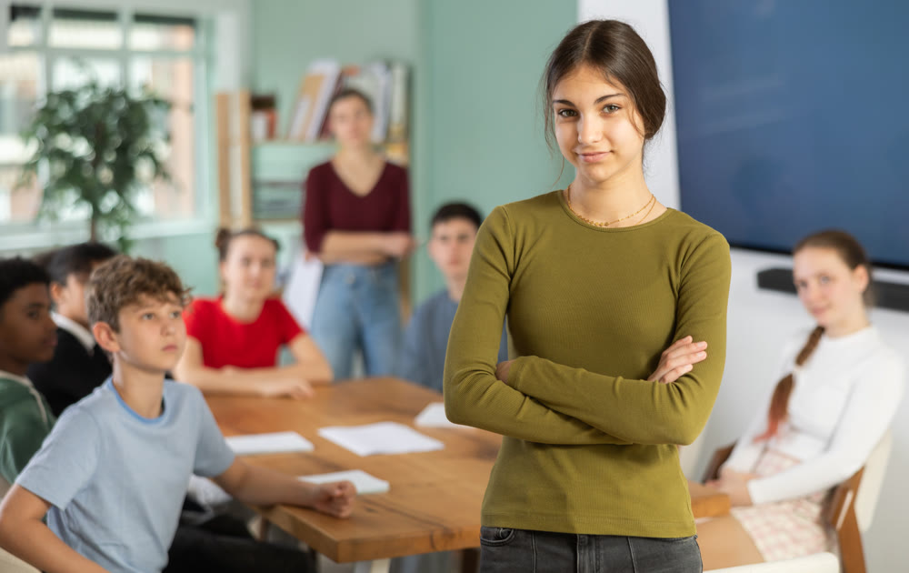 Happy and smiling teen girl stands in classroom, classmates and teacher are behind