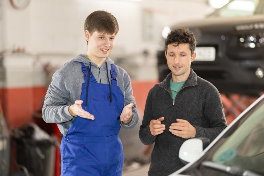 Young man car mechanic talks with man client in workshop and takes order.