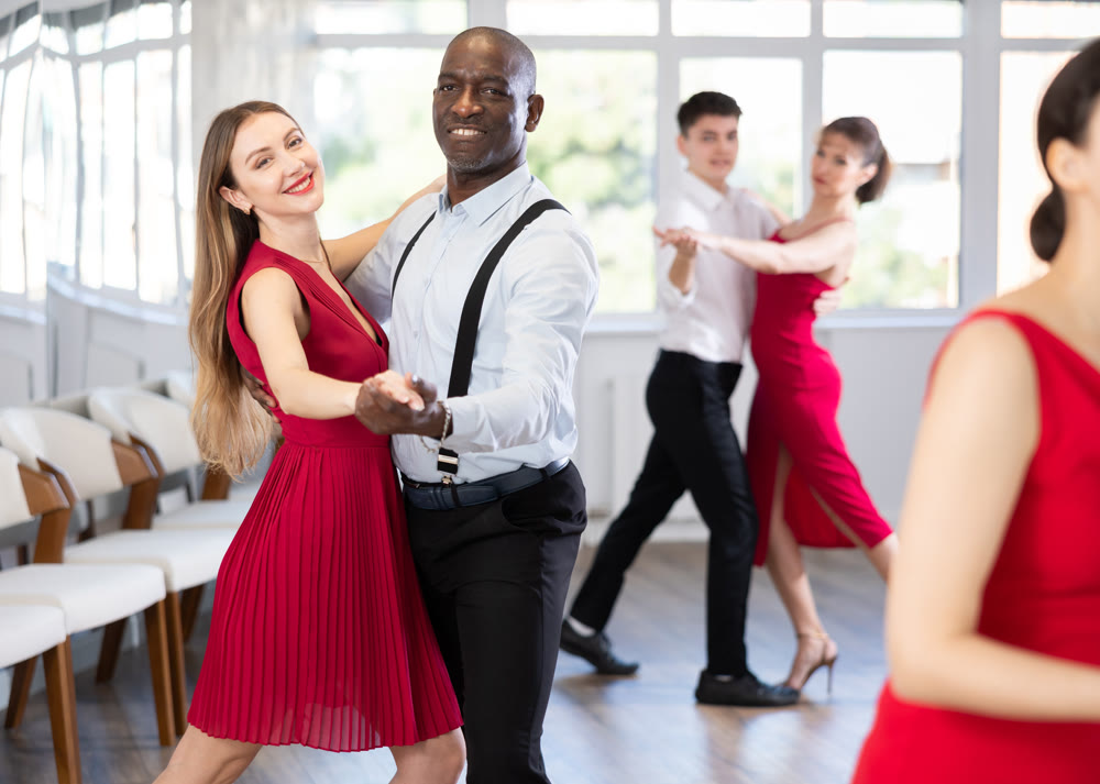 Positive woman in red dress and African american man practicing waltz dance moves in pair during group party in dancing studio