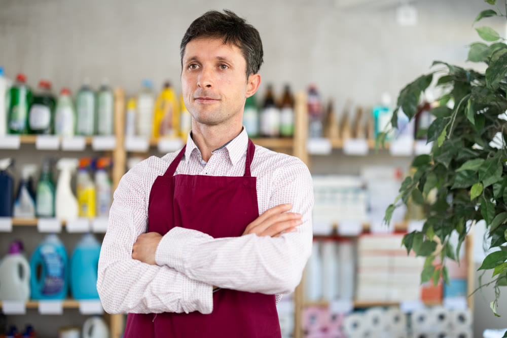 Man in apron stands with arms crossed over chest among shelves and storefronts in household store