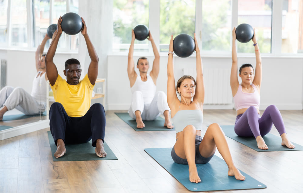 Pilates class in multi-ethnic group - men and women hold the ball above their heads and do bends
